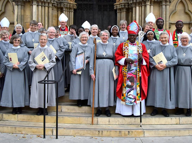 A group of clergy and nuns in gray and white robes pose on church steps. The bishop in the center wears a colorful stole and red robe, exuding joy.