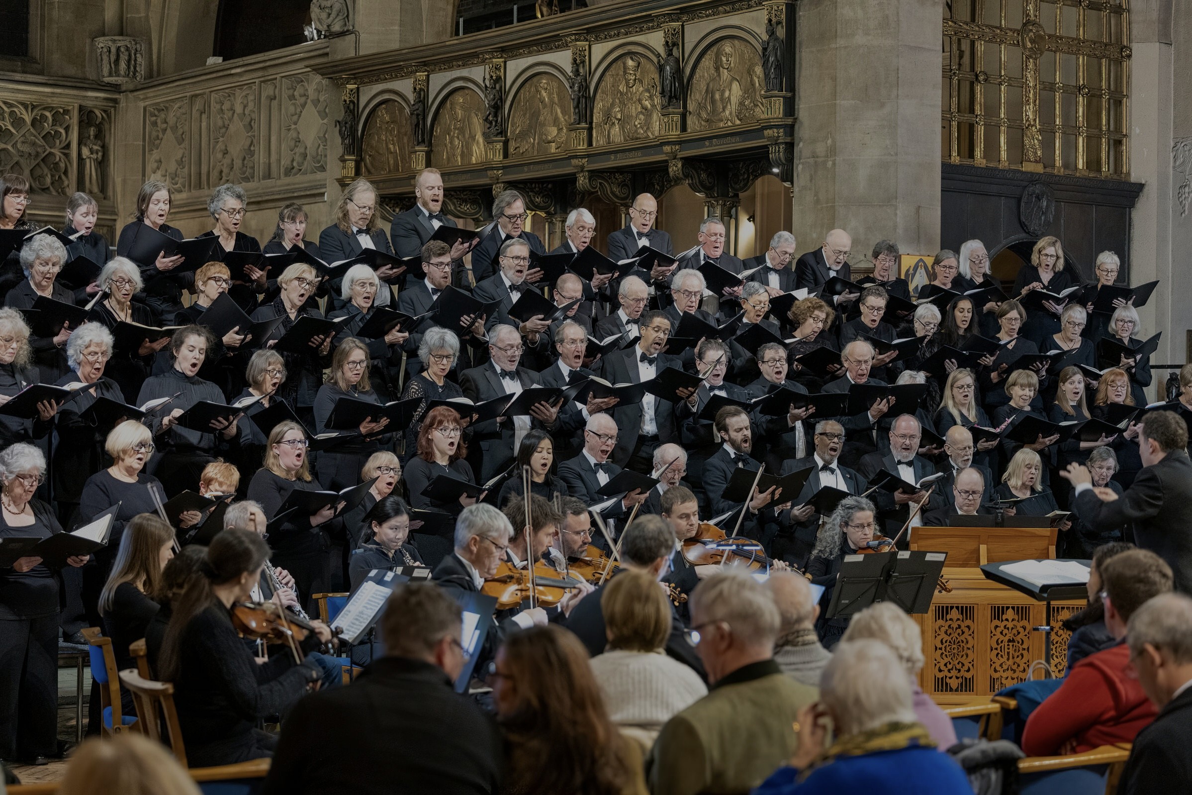 A large choir and orchestra perform in a grand church, with an audience seated in front. The choir members hold black folders and wear black attire. Ornate gold and stone architectural details are visible in the background.