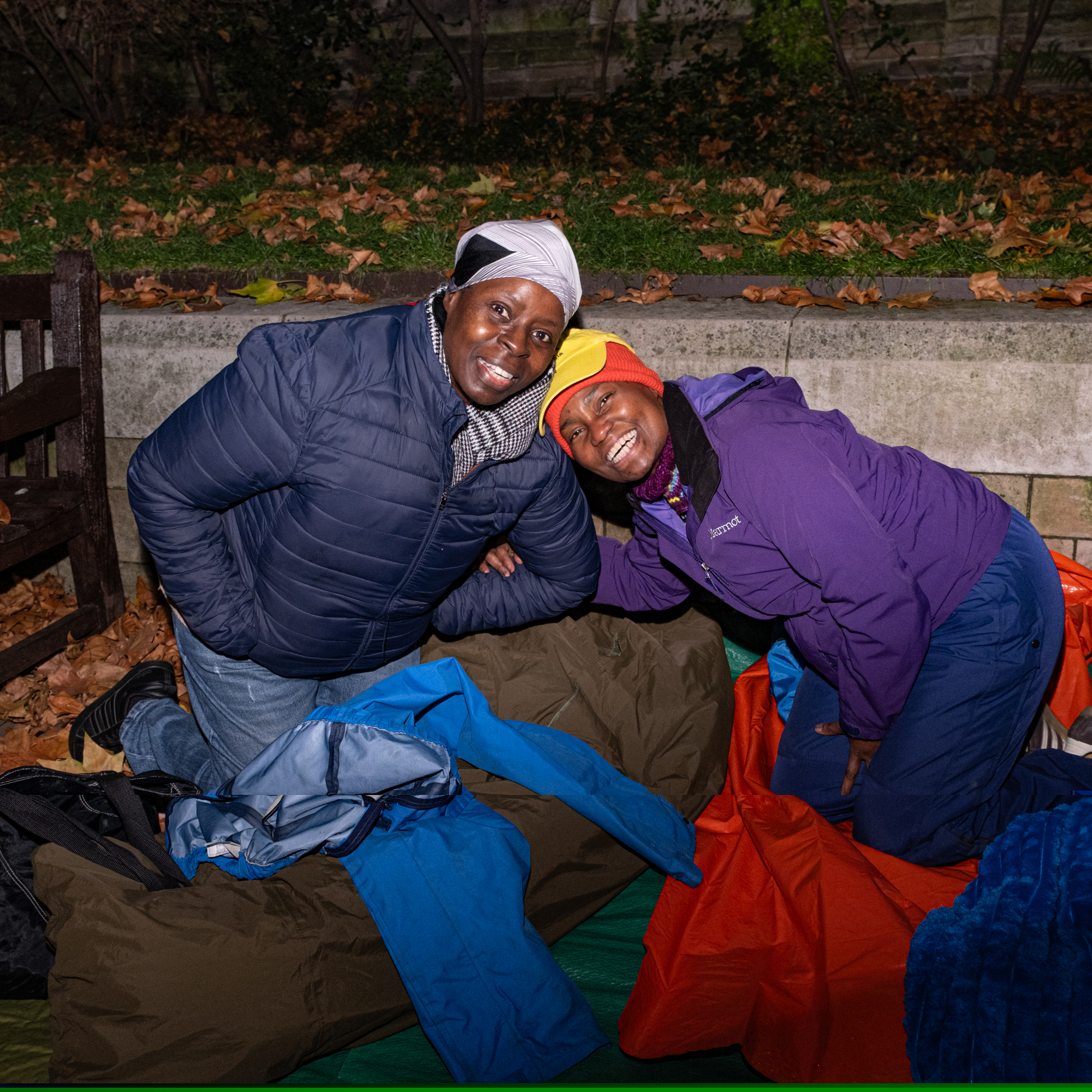 Two smiling women kneeling outdoors at night next to a pile of sleeping bags and outdoor gear.